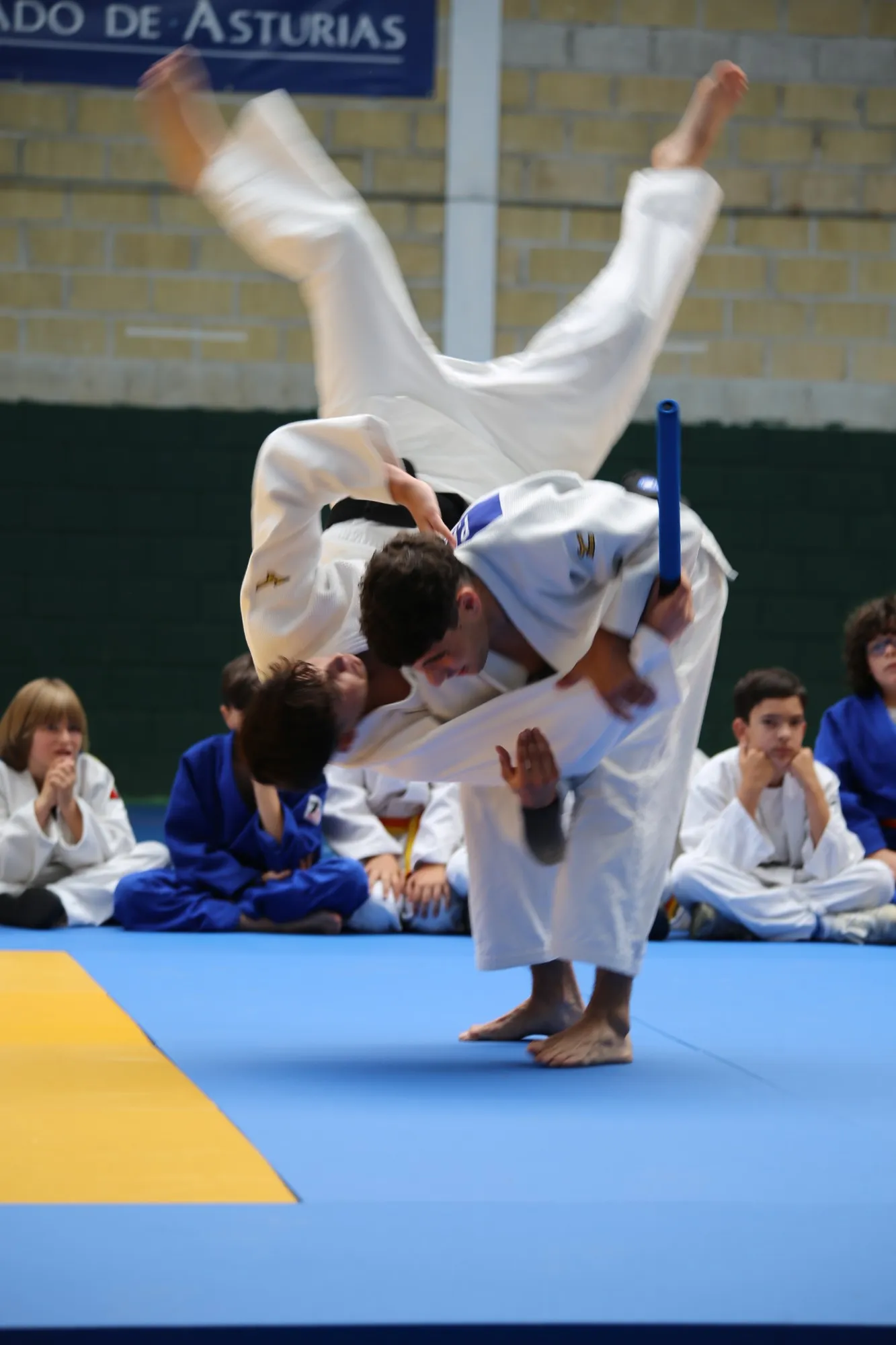 Competición en Nava: un judoka proyecta a otro por encima del hombro mientras los niños miran sentados en el tatami